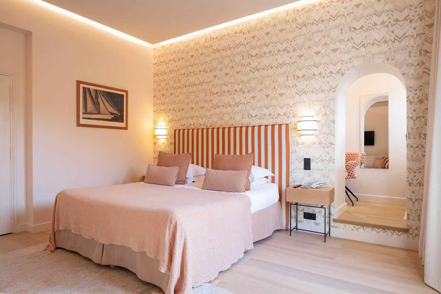Chambre aux tons pêche avec tête de lit rayée orange, couvre-lit rose poudré, papier peint à motifs beige, et arche menant à un salon attenant.