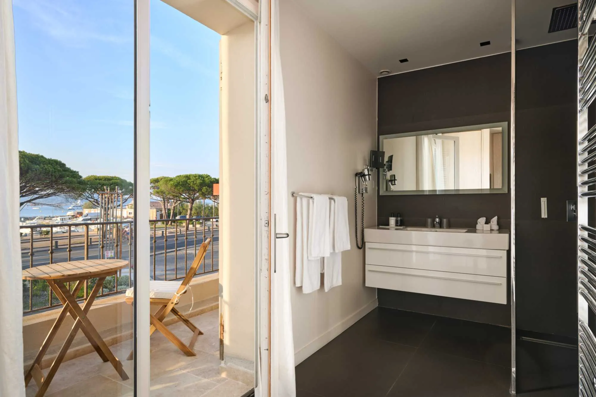 Salle de bain contemporaine ouvrant sur un balcon avec vue sur les pins parasols et le port, vasque blanche suspendue sur mur foncé et douche à l'italienne.