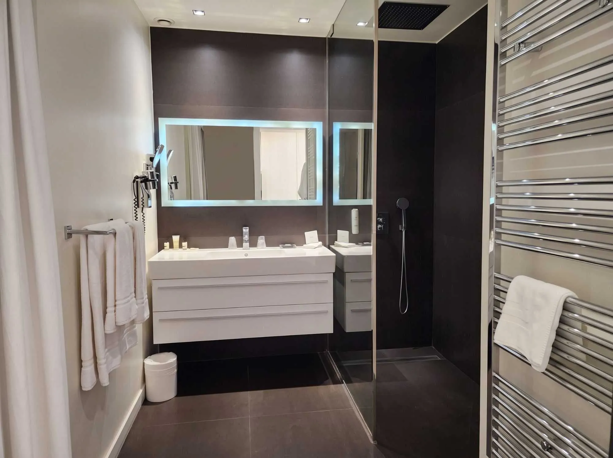 Salle de bain moderne aux tons marron foncé, avec vasque blanche suspendue, miroir rétroéclairé, douche à l'italienne vitrée et sèche-serviettes chromé.
