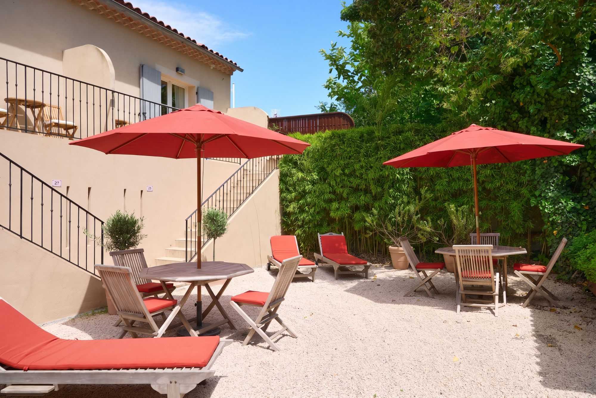 Cour ensoleillée d'une villa provençale avec parasols rouges, chaises longues et tables en bois sur gravier blanc, entourée d'une haie de bambous verts.