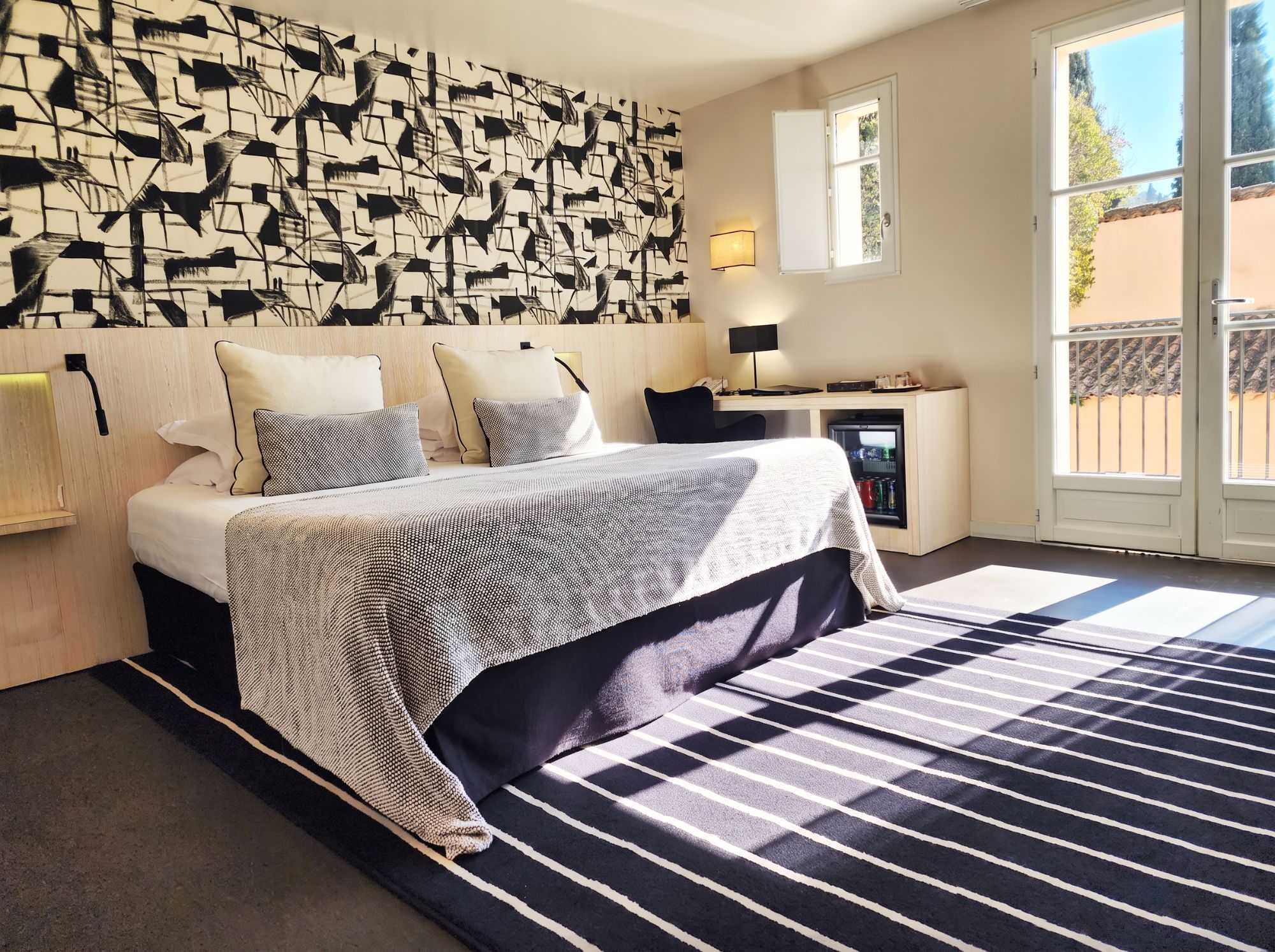 Chambre lumineuse au papier peint graphique noir et blanc, tapis rayé assorti, minibar et bureau, avec porte-fenêtre ouvrant sur un balcon baigné de soleil.