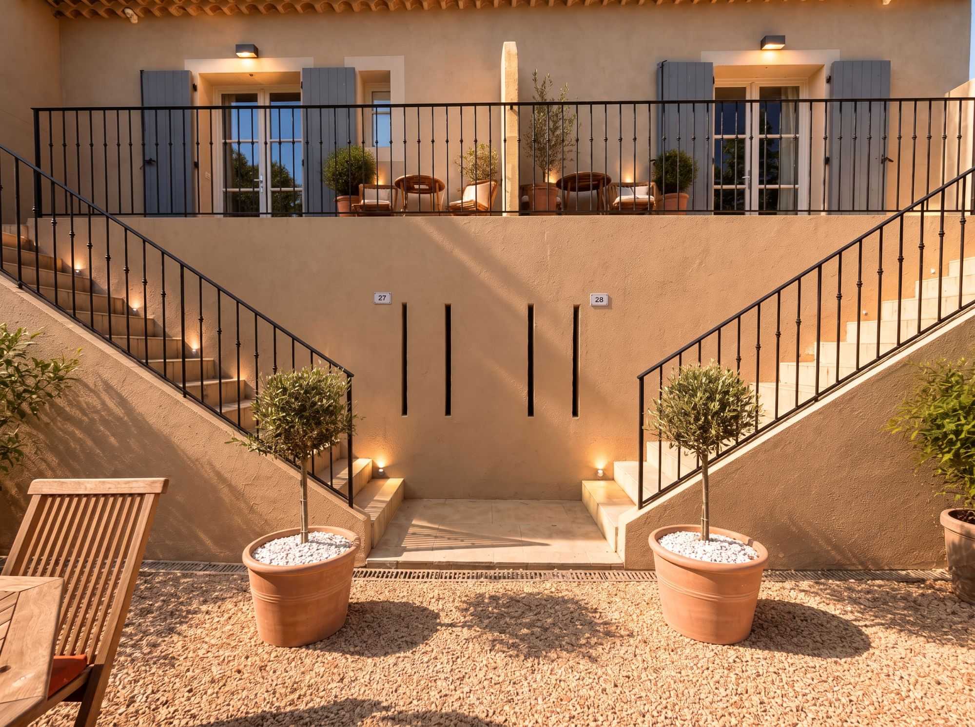 Cour symétrique d'une villa aux volets bleu-gris, avec deux escaliers en fer forgé encadrant l'entrée, oliviers taillés en pots et terrasse aménagée à l'étage.