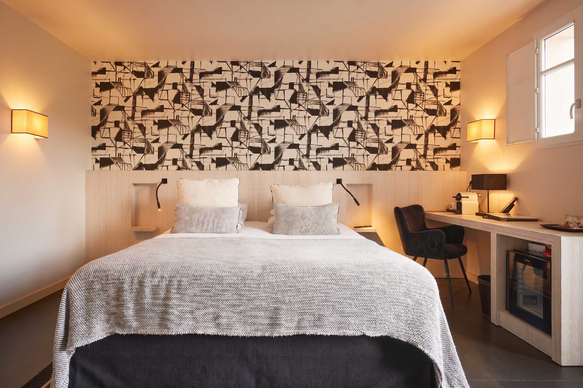 Chambre moderne avec papier peint graphique noir et blanc au-dessus du lit, appliques lumineuses intégrées à la tête de lit, et bureau avec fauteuil noir et minibar.