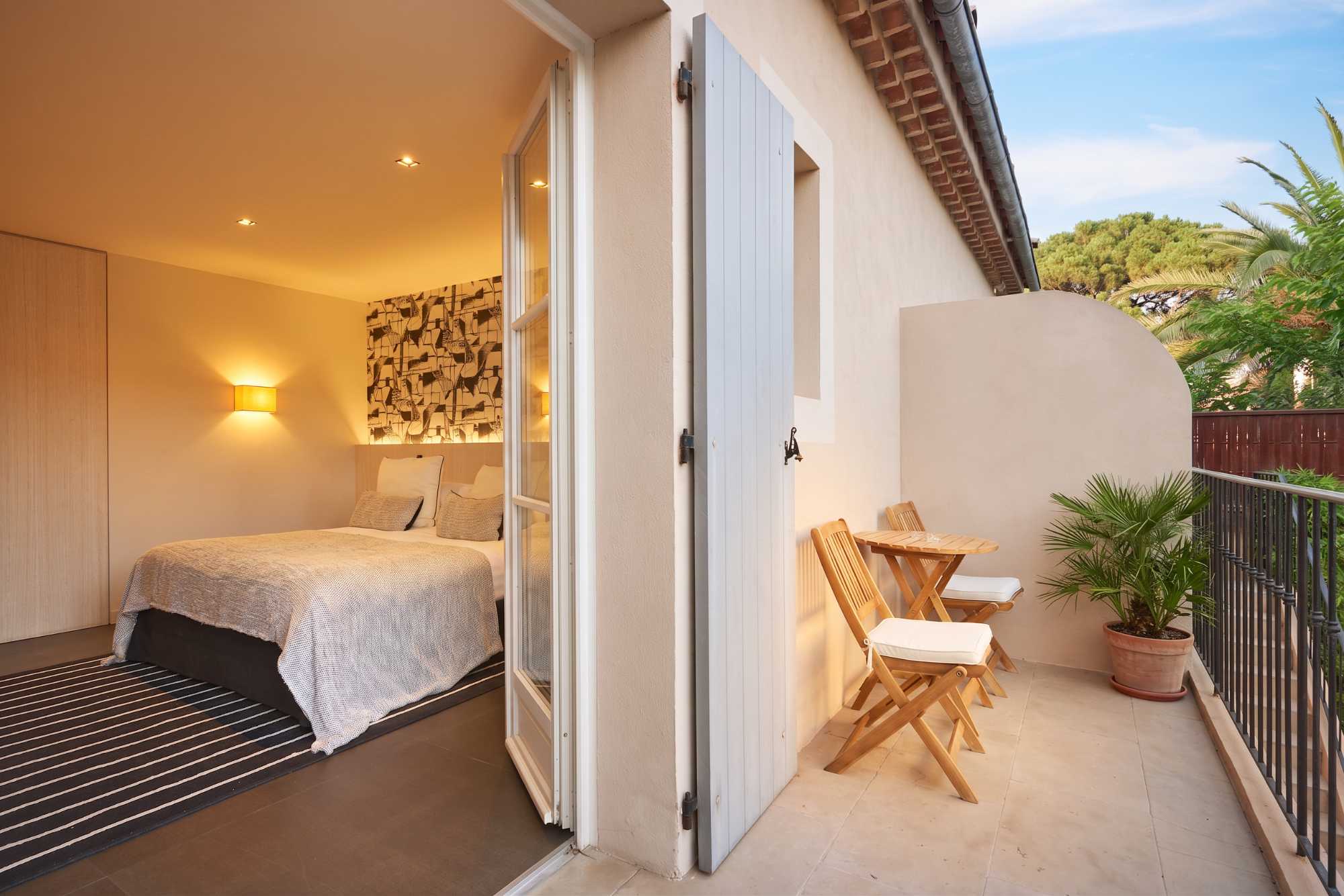 Chambre chaleureuse s'ouvrant sur un balcon privé avec table et chaises pliantes en bois, palmier en pot, et vue sur des pins et un mur en pierre.