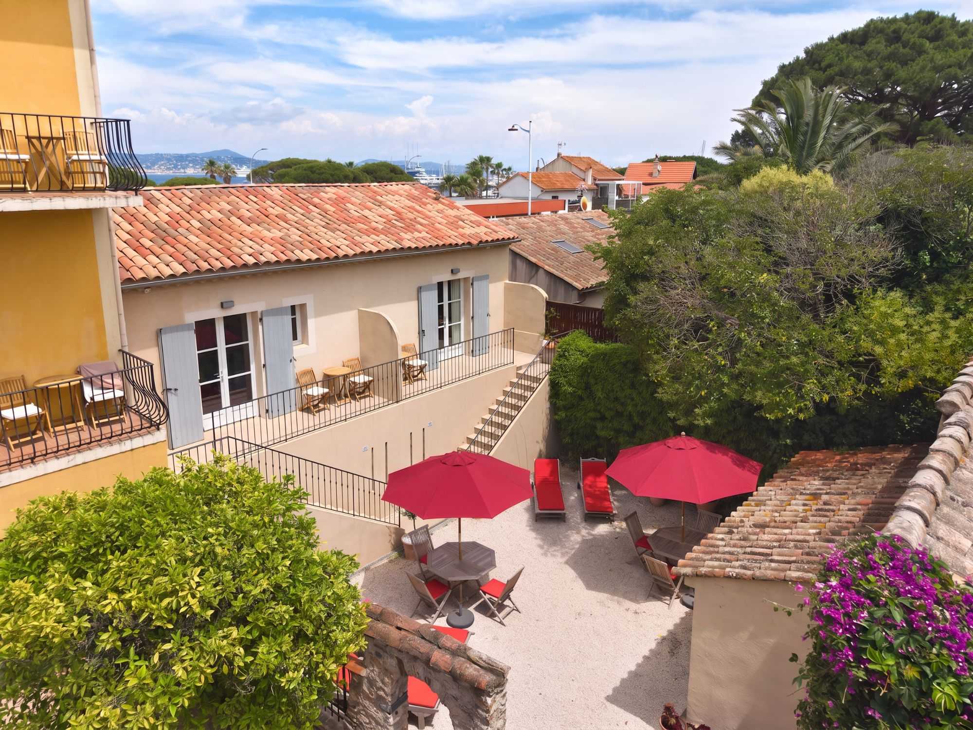 Vue en hauteur d'une terrasse méditerranéenne avec parasols rouges, transats et tables, entourée de toits en tuiles, verdure luxuriante et vue lointaine sur le port.