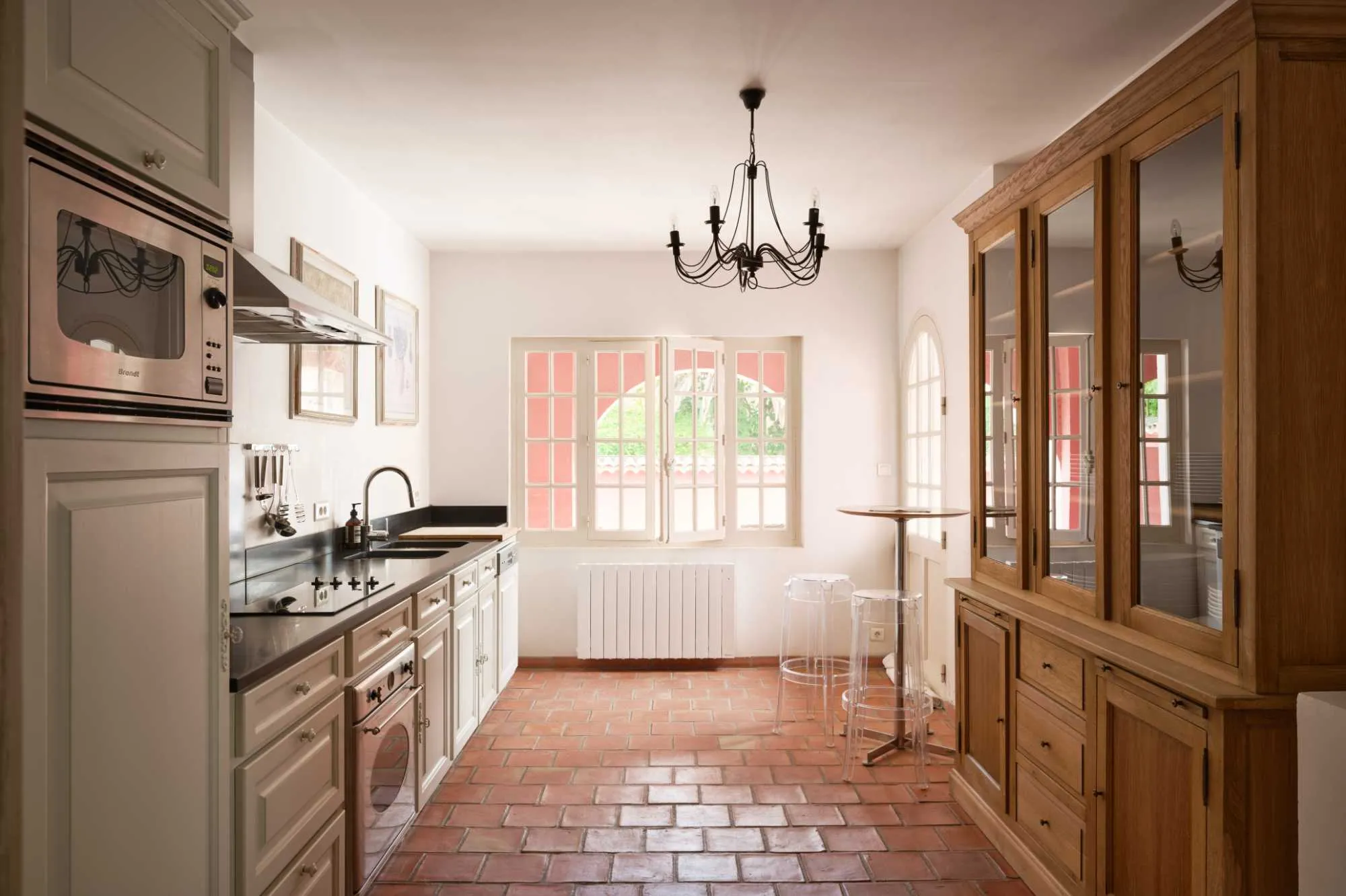 Cuisine provençale au sol en tomettes, avec buffet vitré en bois, plan de travail noir, lustre en fer forgé, et fenêtres à petits carreaux laissant entrer la lumière.