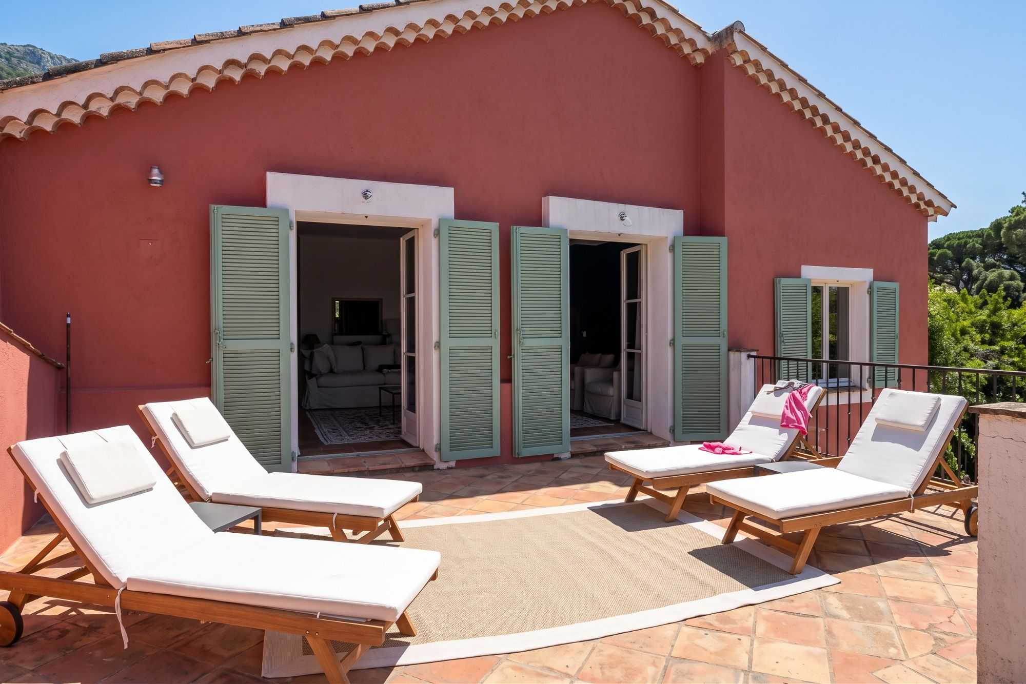 Terrasse ensoleillée d'une villa aux murs terracotta et volets verts, avec quatre chaises longues blanches disposées devant des portes-fenêtres ouvertes.