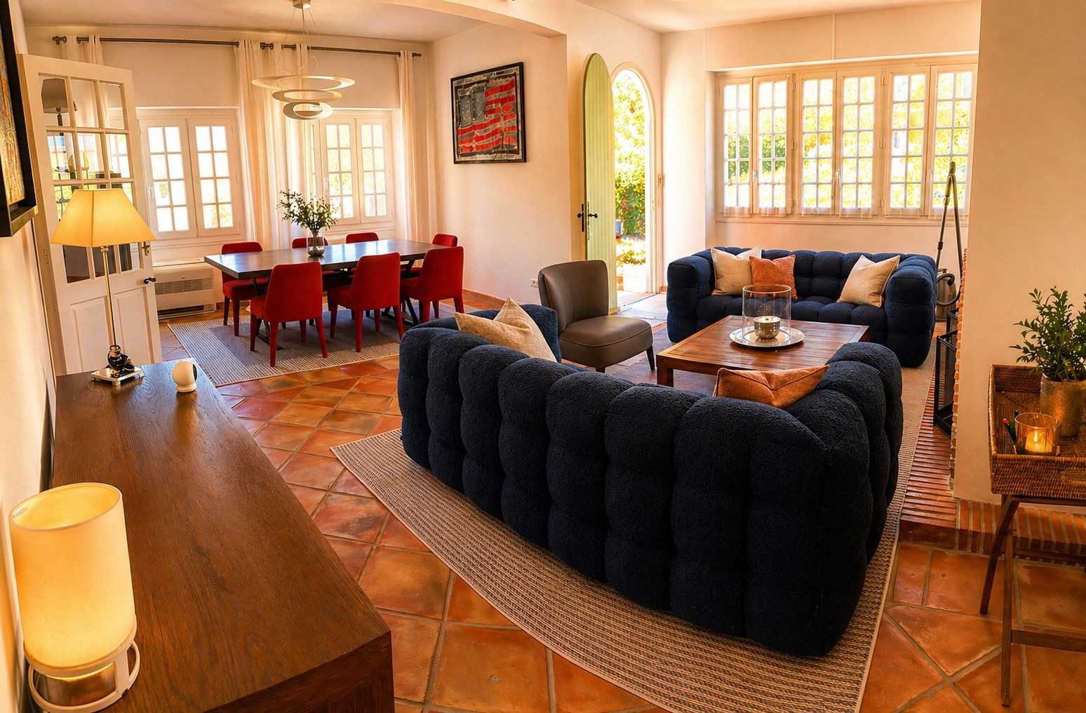 Salon-salle à manger provençal au sol en terre cuite, avec canapés matelassés bleu marine, table à manger et chaises rouges, et grandes fenêtres à petits carreaux baignées de lumière.