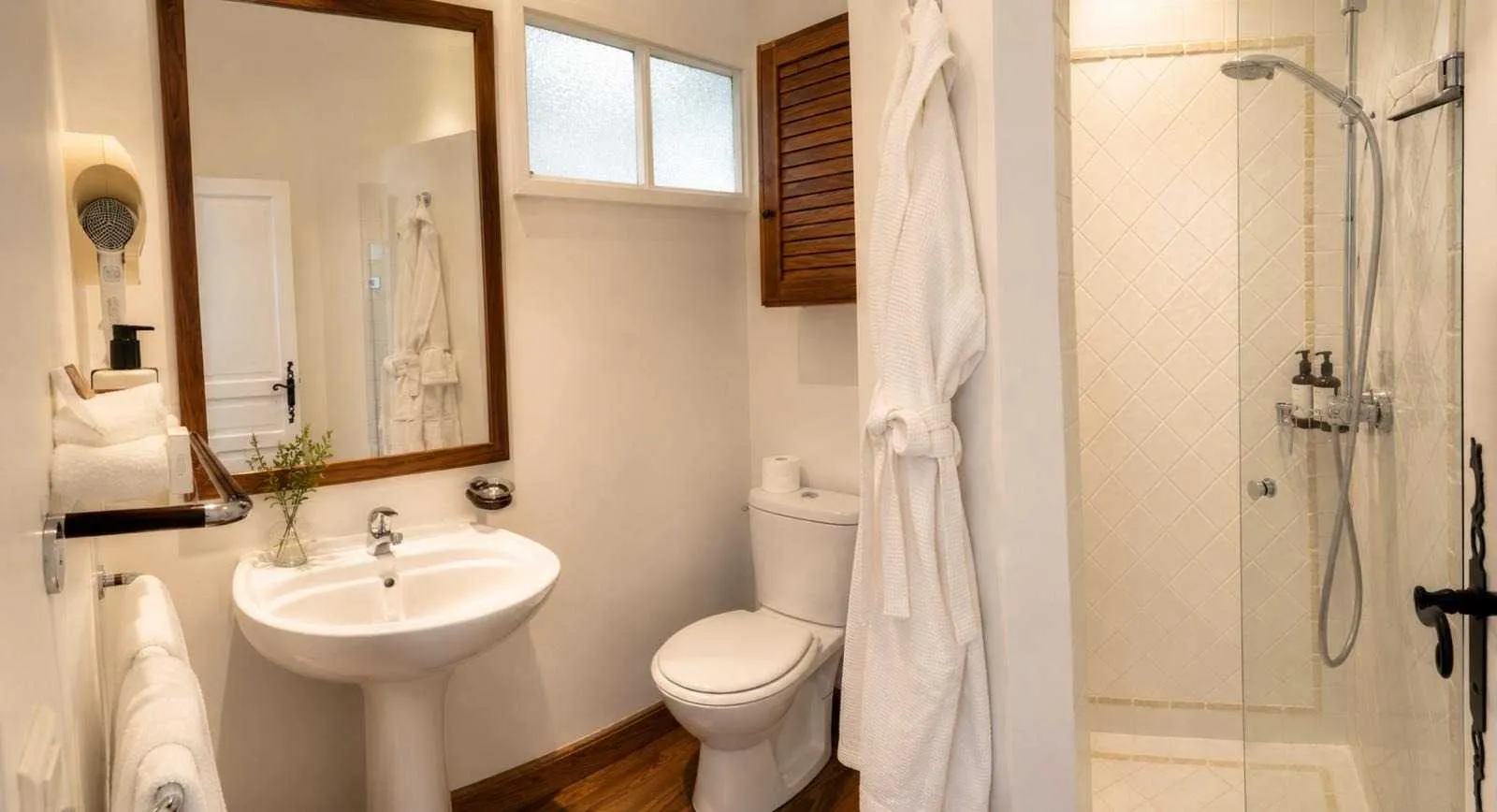 Petite salle de bain claire avec lavabo sur pied, WC, peignoir blanc suspendu, et douche vitrée aux carreaux beige en losange.