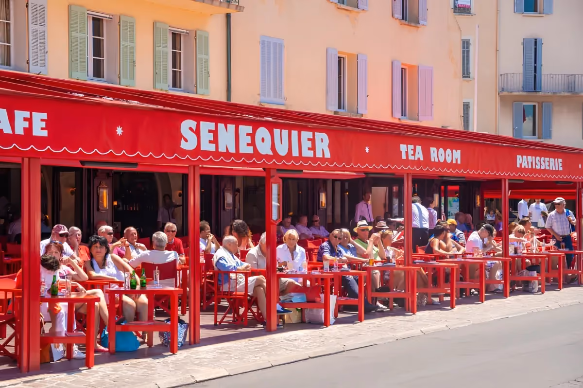 Terrasse animée du célèbre café Sénéquier à Saint-Tropez, avec sa façade et son mobilier rouge emblématique, des clients installés devant des façades pastel aux volets colorés.