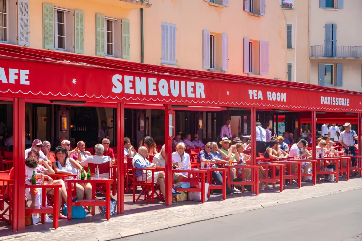 Terrasse animée du célèbre café Sénéquier à Saint-Tropez, avec sa façade et son mobilier rouge emblématique, des clients installés devant des façades pastel aux volets colorés.