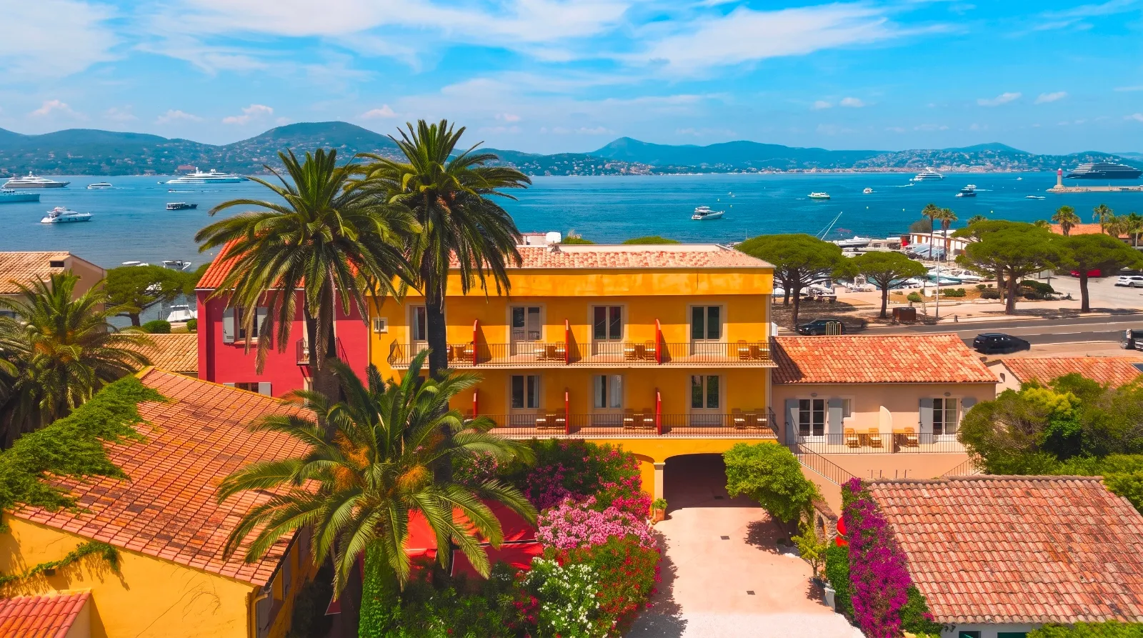 Veduta aerea di un hotel giallo in stile mediterraneo con palme e bouganville, tetti in tegole arancioni, affacciato su una baia con yacht e colline sullo sfondo.