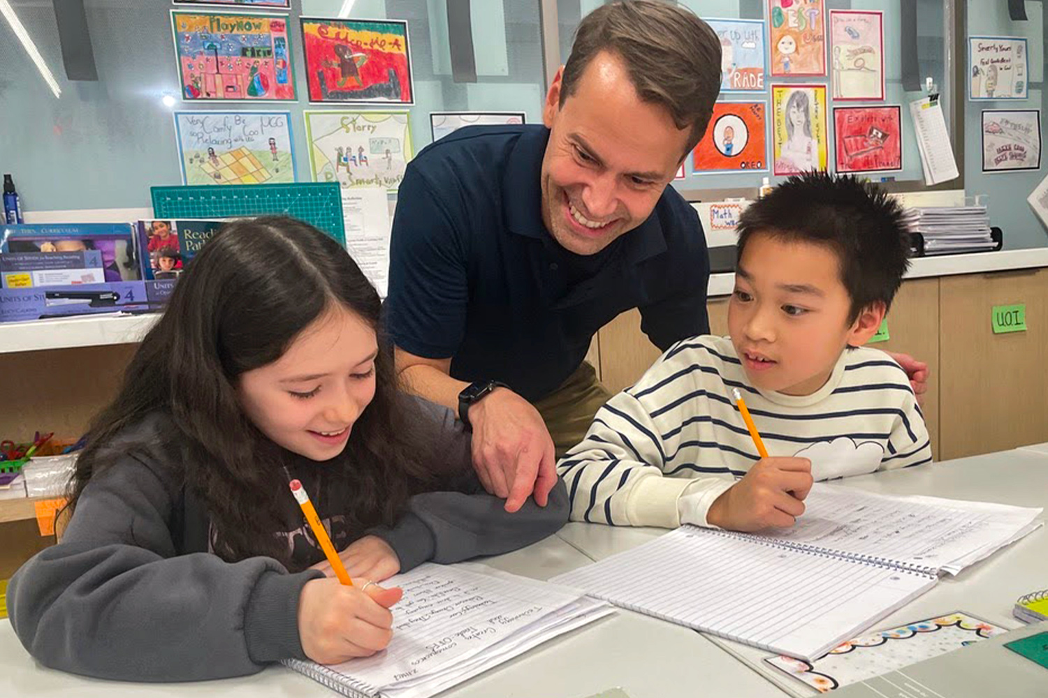 Two Pine Street School students are working at a desk and their teacher is pointing out something on one student's notebook.