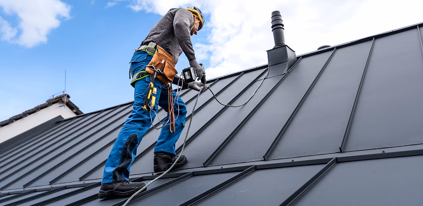 Ein Handwerker mit Helm und Werkzeugfach arbeitet auf einem Metall-Dach bei bewölktem Himmel.