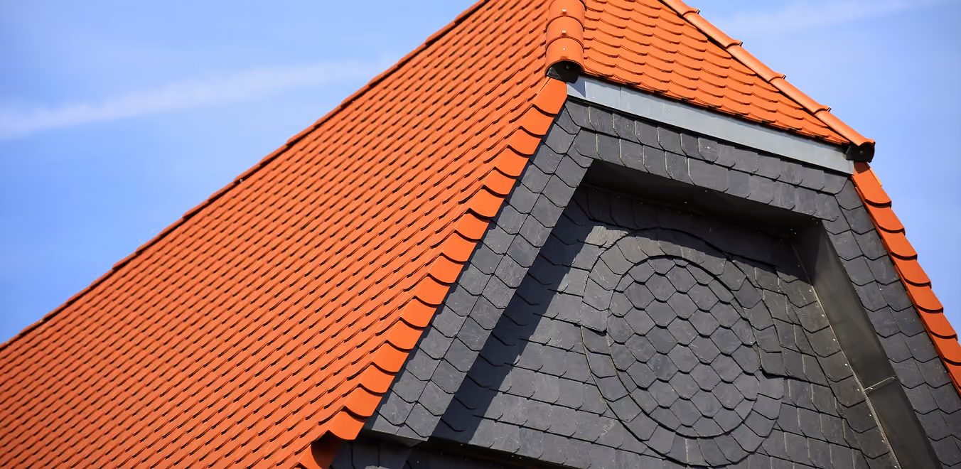 Nahaufnahme eines roten Ziegeldachs mit grauem Schieferdetail und blauem Himmel im Hintergrund.