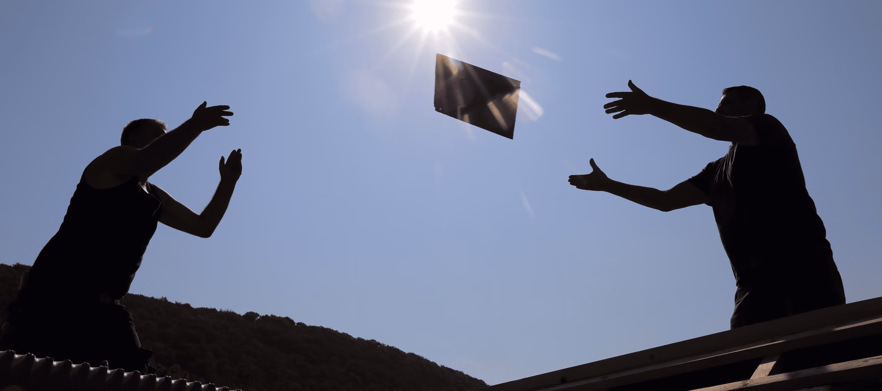 Zwei Männer werfen ein rechteckiges Objekt zueinander vor dem hellen Himmel mit Sonnenstrahlen.
