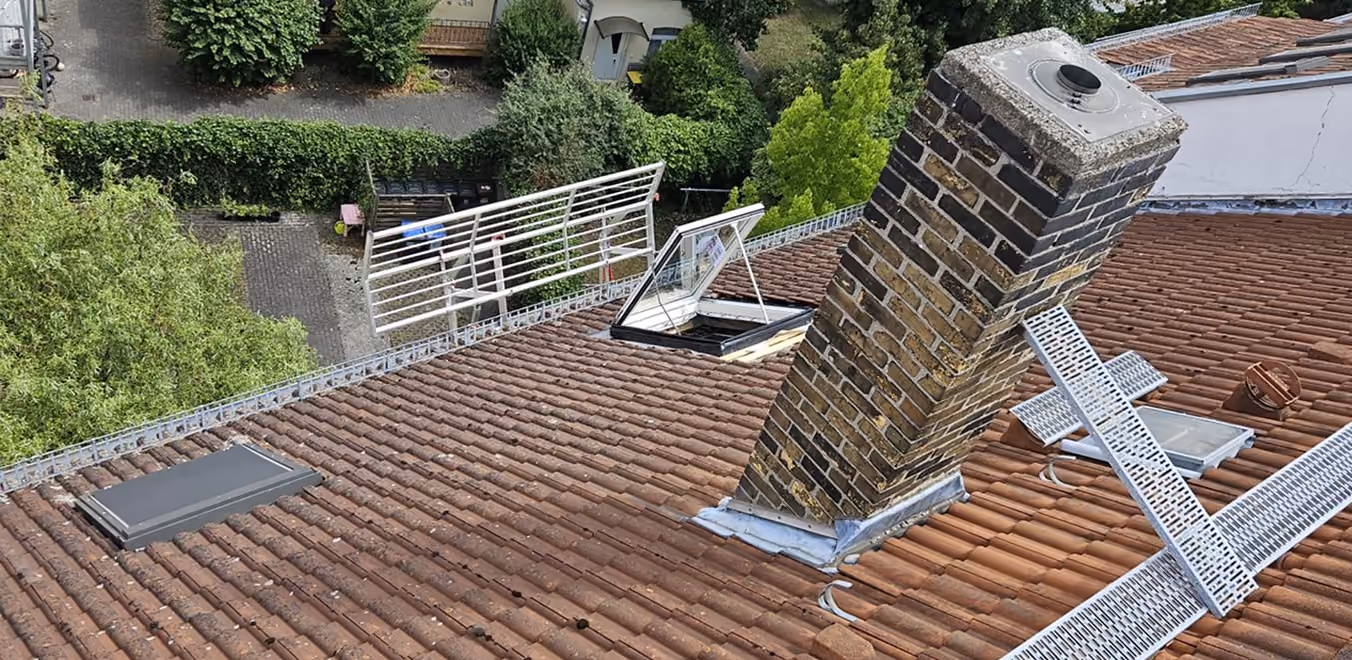 Dach mit Kamin, offenem Dachfenster, Dachrinnenrinnen und Bäumen im Hintergrund.
