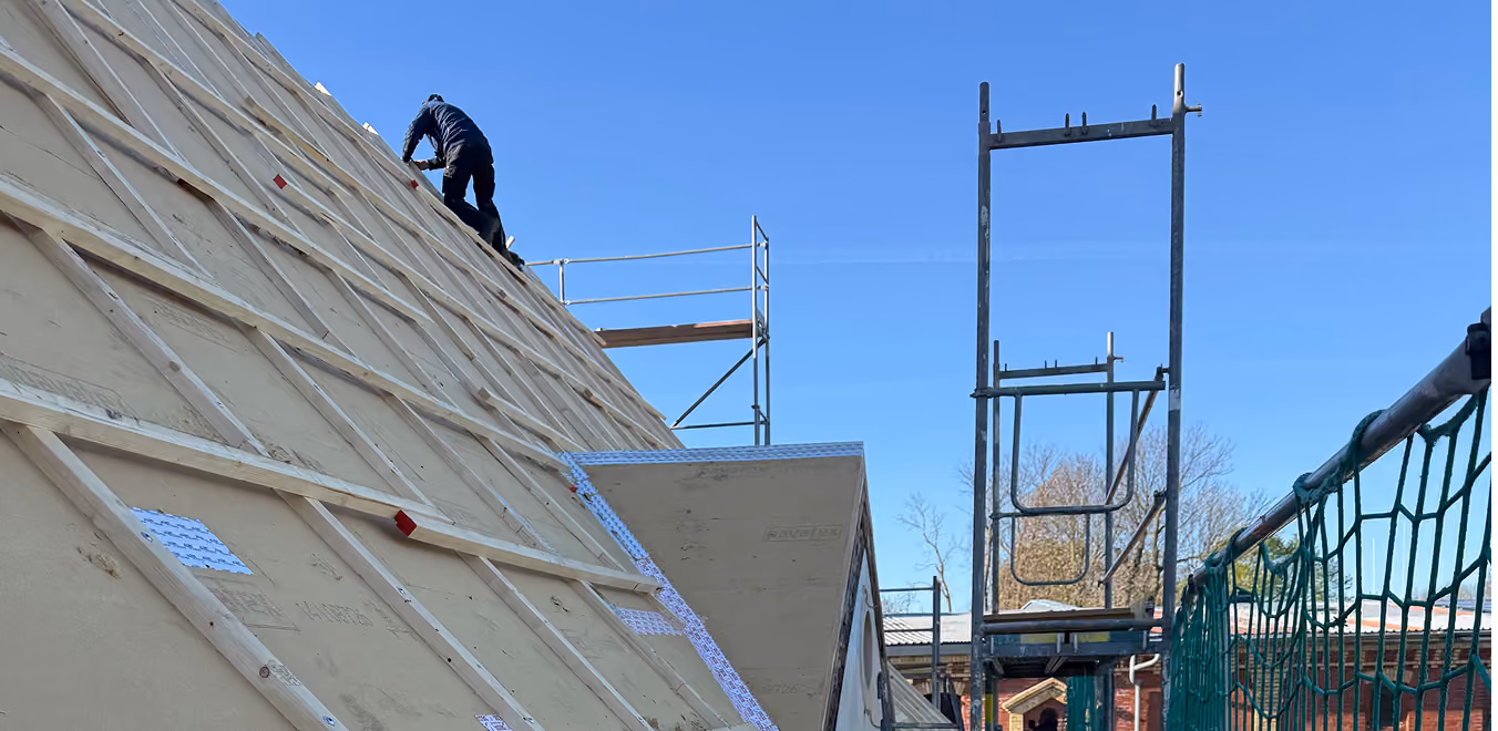 Bauarbeiter arbeitet am geneigten Dach mit Holzrahmen und Gerüst bei klarem Himmel.