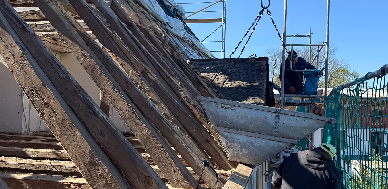 Dachdecker arbeiten an einem schrägen Dach mit Holzstützen und Hebekorb bei klarem Himmel.