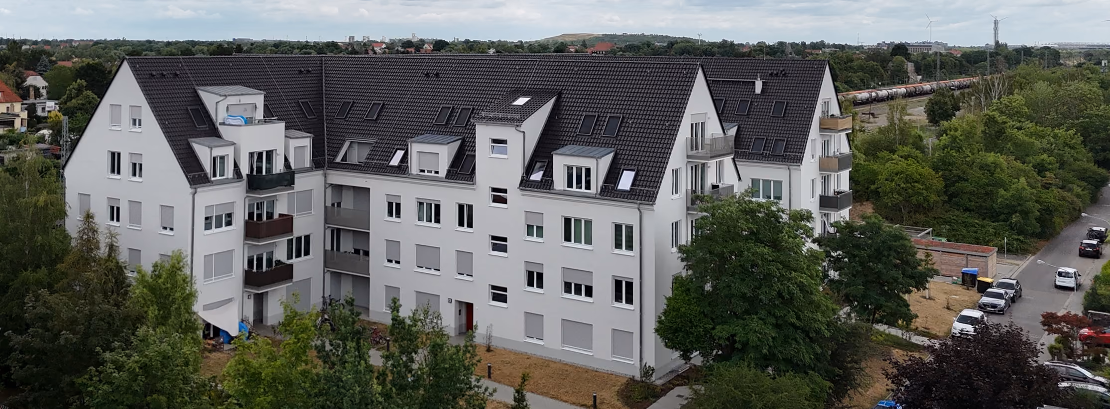 Mehrstöckiges Wohngebäude mit dunklem Dach, Balkonen und umliegenden Bäumen an einer Straße.