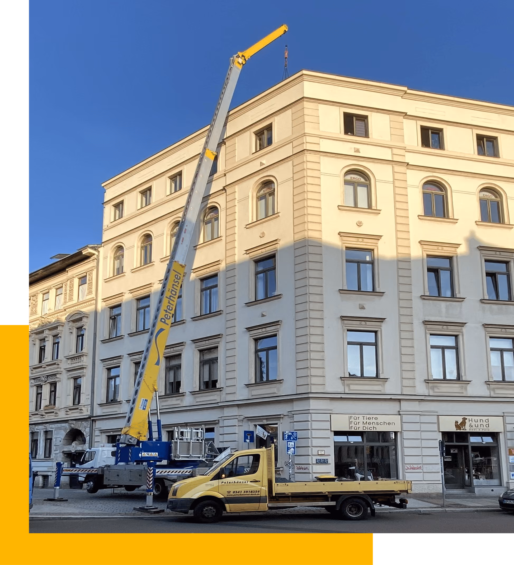 Gelber Kran hebt Last an der Ecke eines mehrstöckigen Gebäudes bei blauem Himmel.