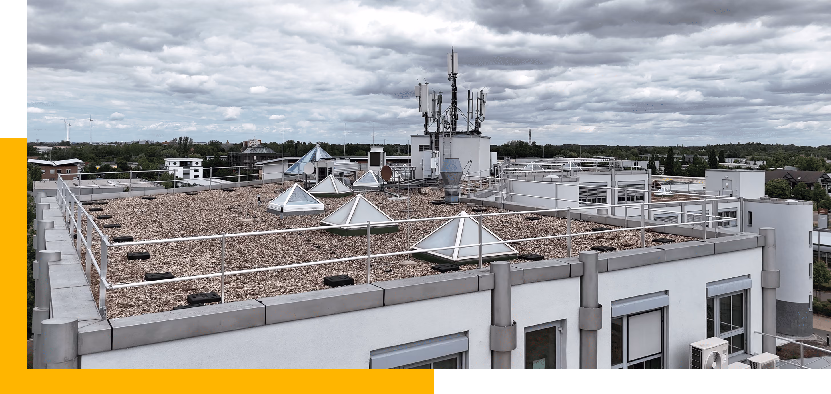 Flachdachgebäude mit metallischem Geländer, pyramidalen Dachfenstern und mehreren Antennen unter bewölktem Himmel.
