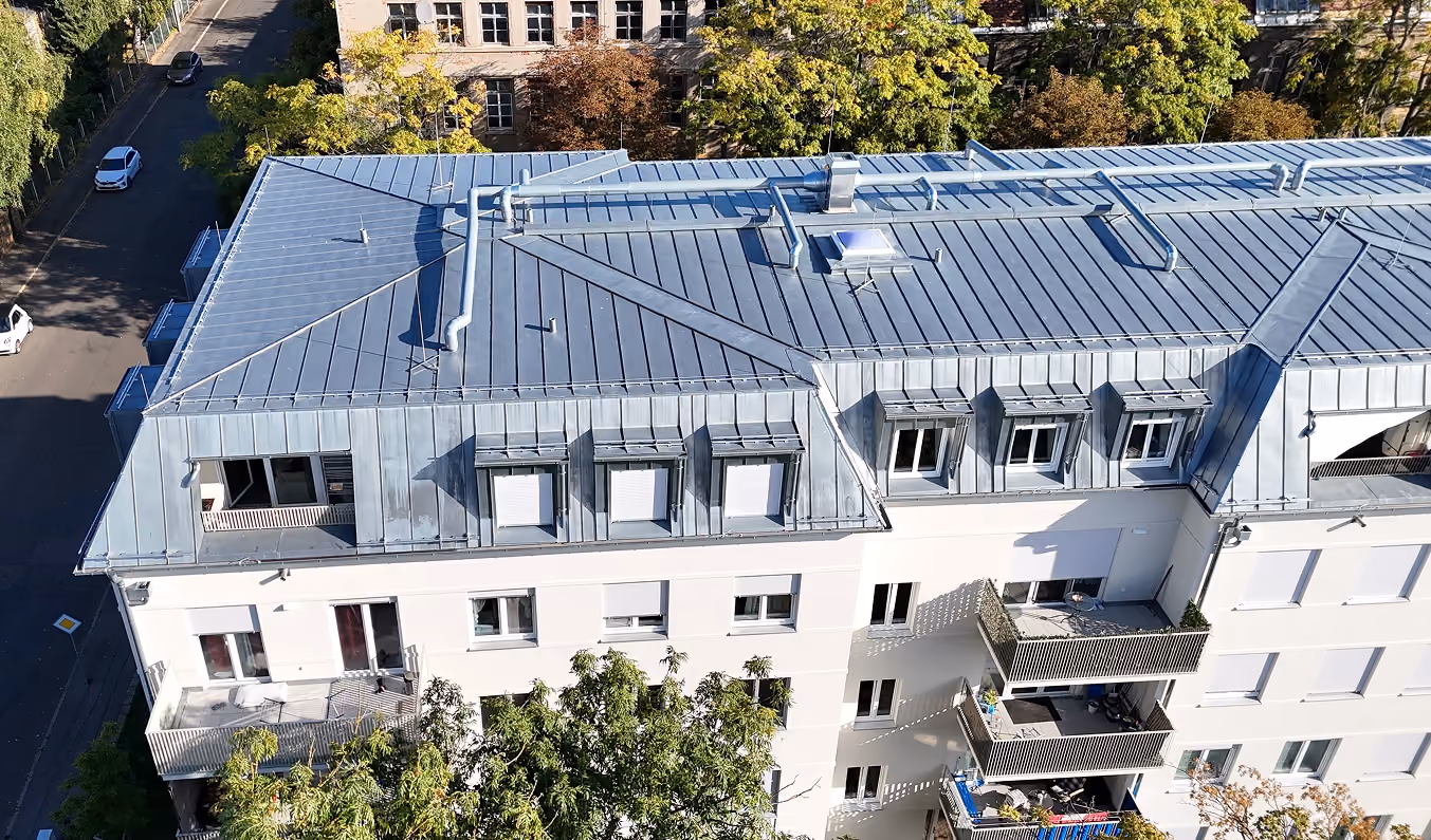Dach eines weißen Wohnhauses mit blauen Fensterläden und mehreren Balkonen, Bäume im Vordergrund.