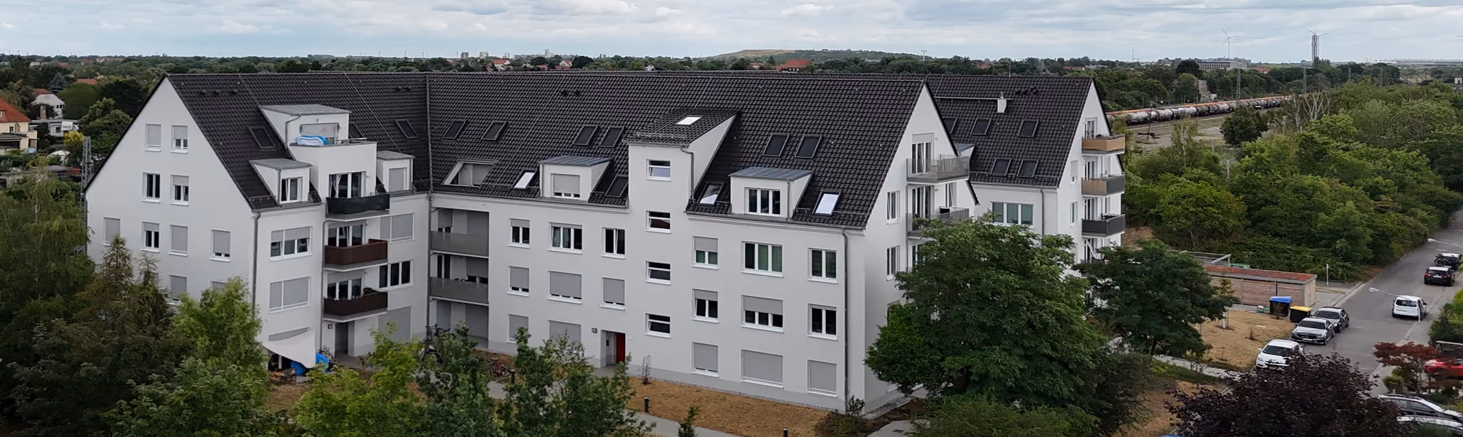 Mehrstöckiges Wohngebäude mit dunklem Dach, weißen Wänden, Balkonen, Bäumen und parkenden Autos.