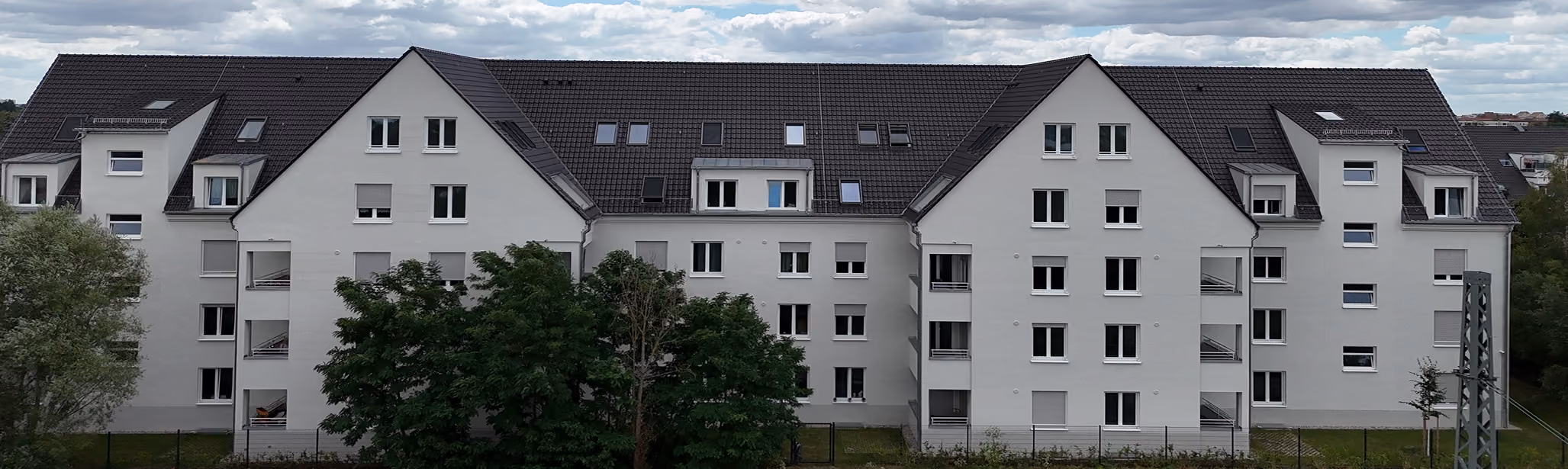 Mehrstöckiges weißes Wohngebäude mit vielen Fenstern und einem dunklen Schrägdach unter bewölktem Himmel.