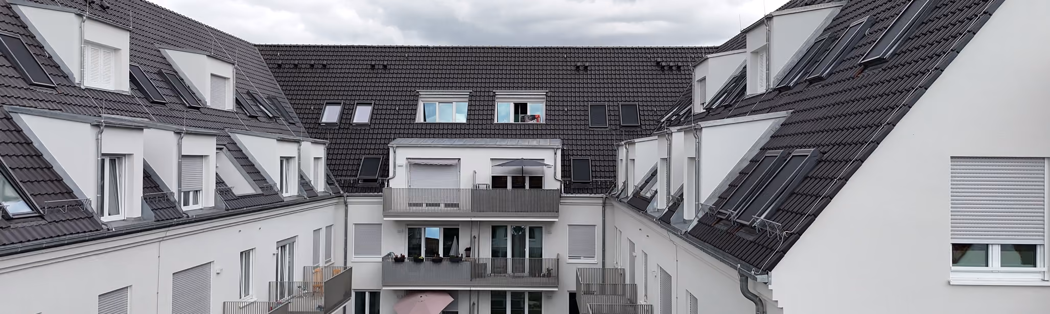 Moderne weiße Wohngebäude um einen Innenhof mit Balkonen und grauen Dächern unter bewölktem Himmel.