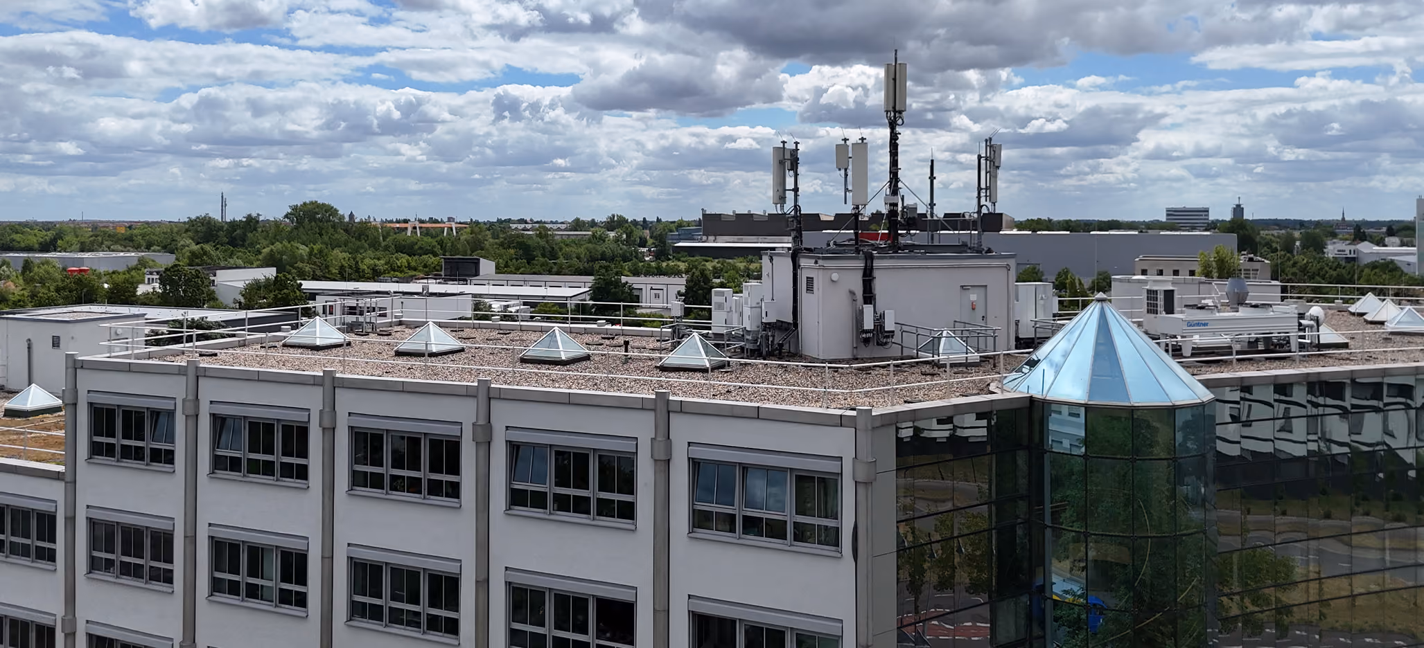 Flachdach eines Bürogebäudes mit Antennen, Dachfenstern und bewölktem Himmel im Hintergrund.