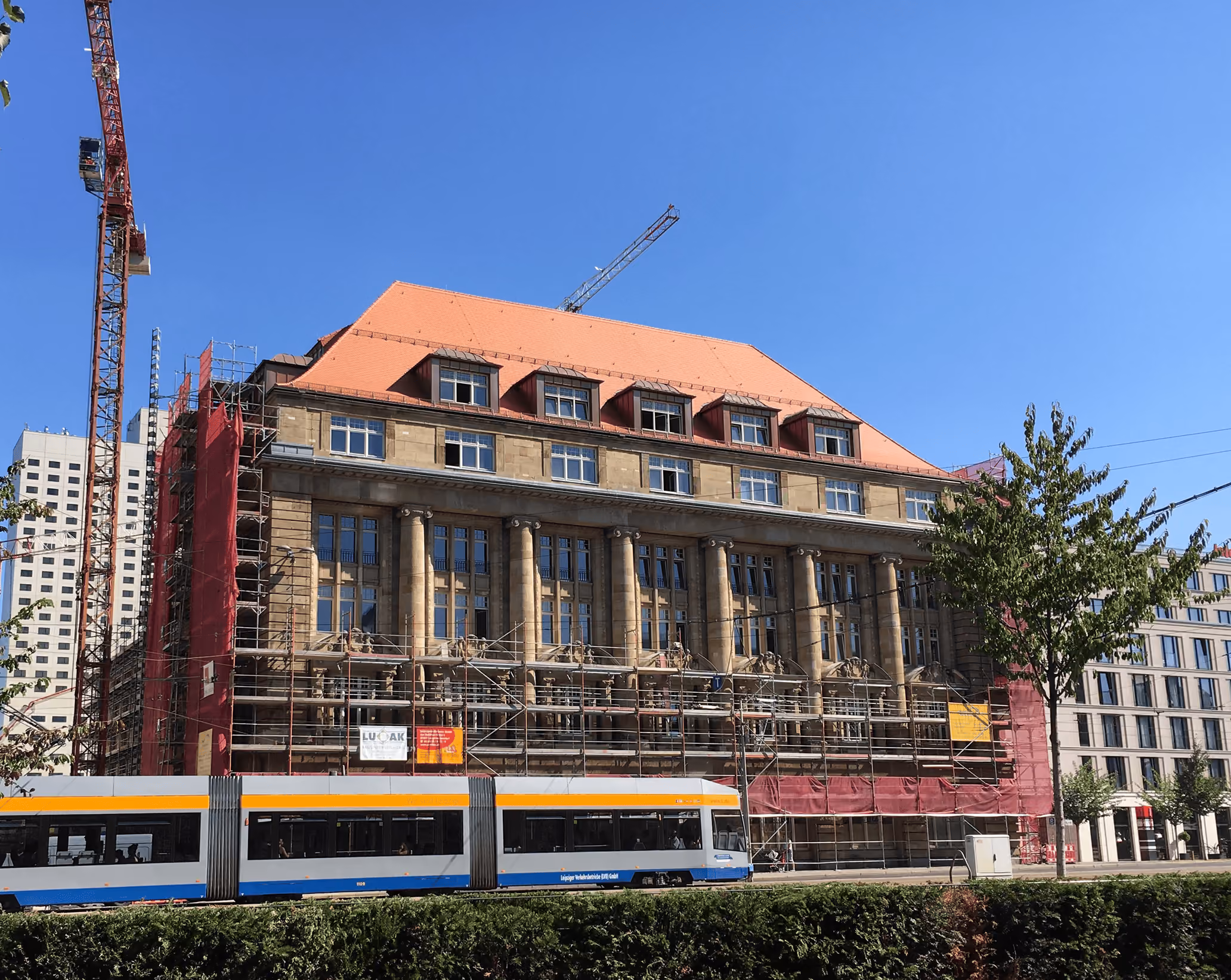 Historisches Gebäude mit roten Dach und Säulen unter Renovierung neben Straßenbahn in klarem Himmel.