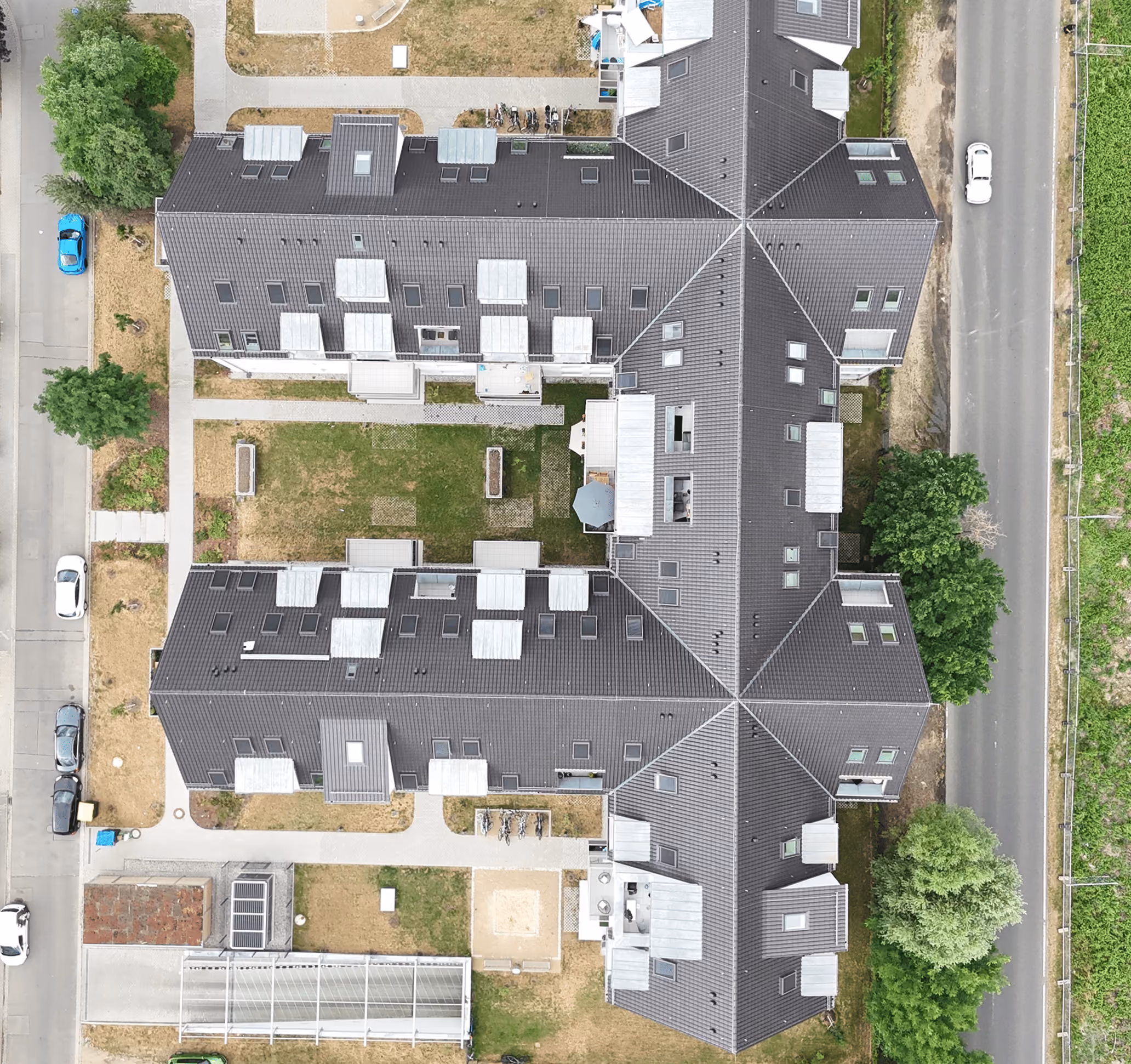 Luftaufnahme eines Gebäudekomplexes mit dunklen Dächern und umgeben von Straßen und Bäumen.