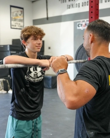 Young man listens to a coach while leaning on a barbell during a beginner fitness class.