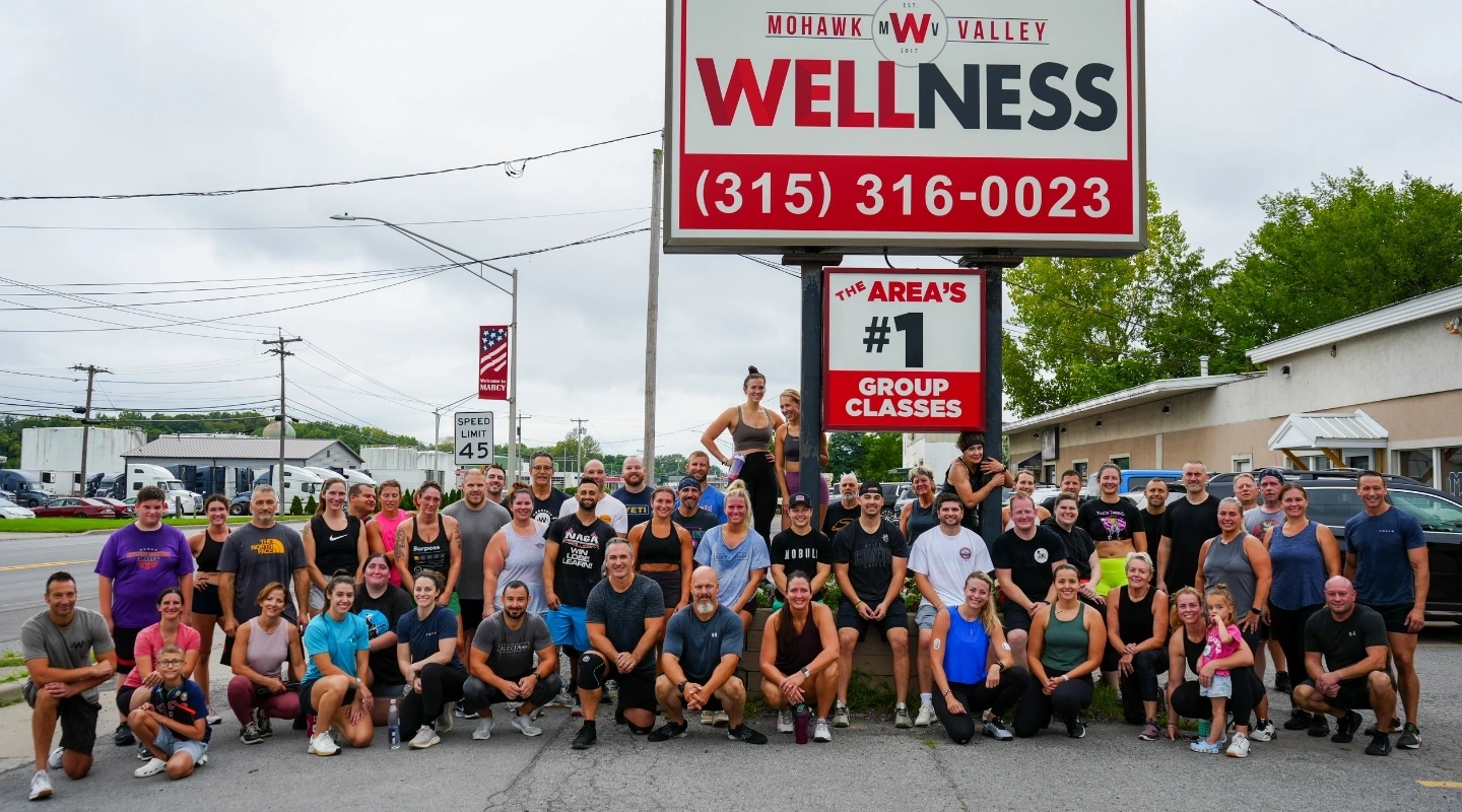 A large group of smiling fitness participants poses outdoors in front of a Mohawk Valley Wellness sign.