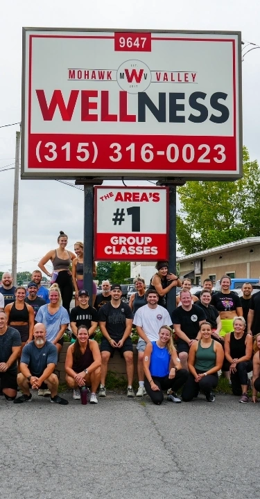 A large group of smiling fitness participants poses outdoors in front of a Mohawk Valley Wellness sign.