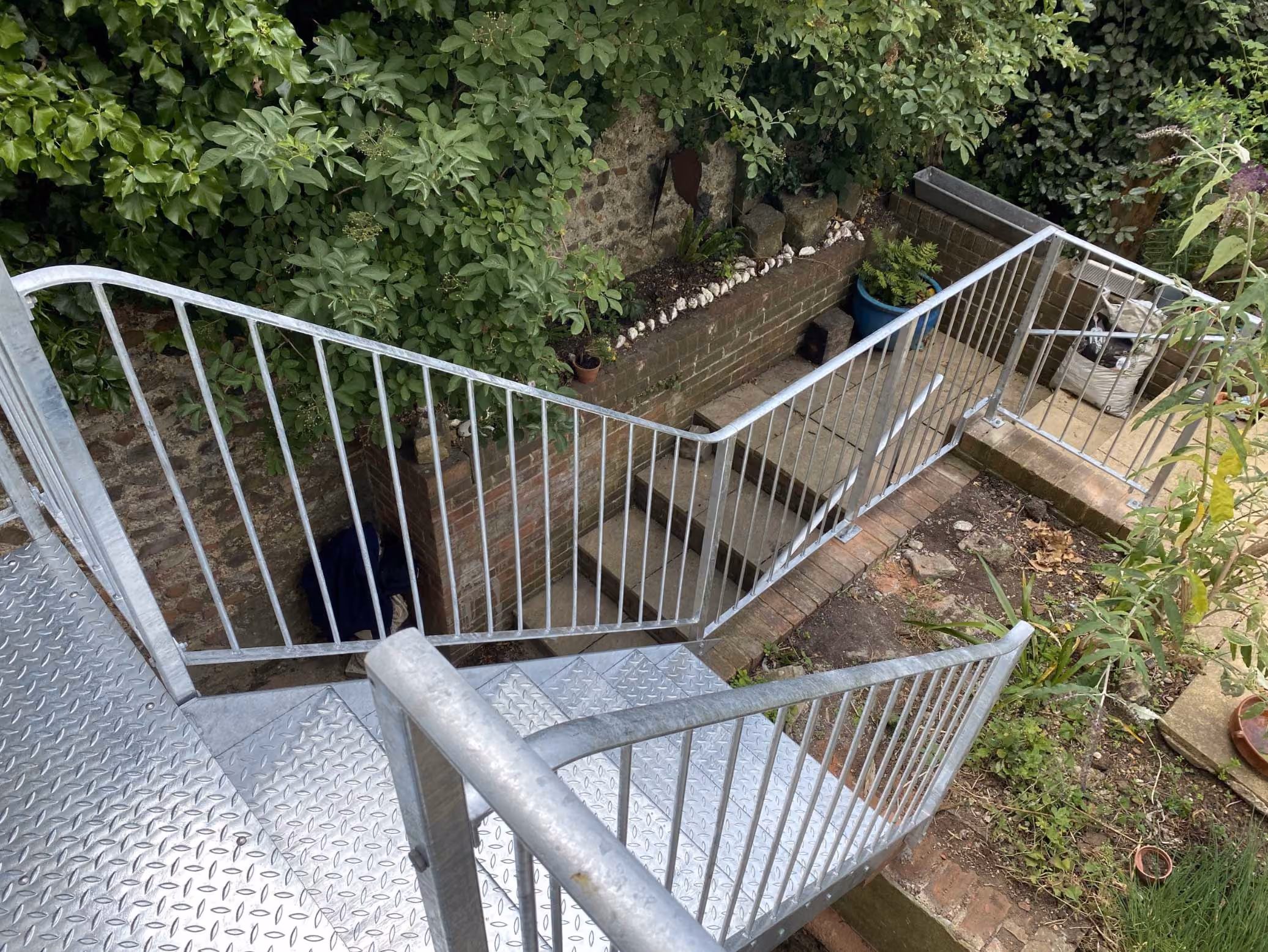 Metal staircase leading down to a garden area with green plants and brick walls.