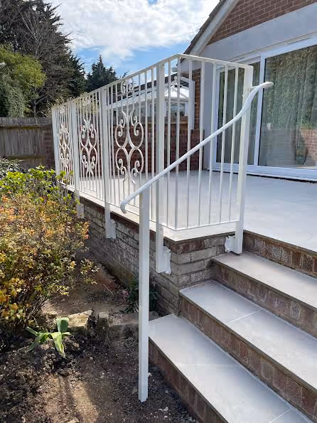White metal railing with decorative patterns on a raised patio, next to brick steps and shrubs.