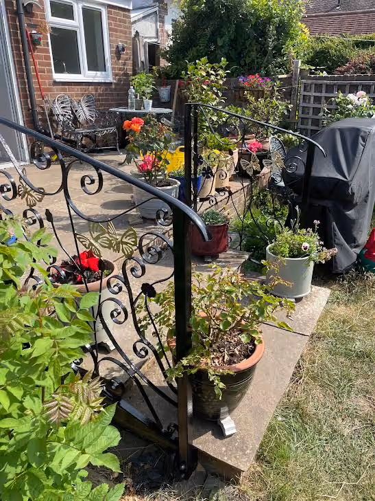 A garden patio with potted flowers, ornate metal railings, and nearby garden chairs in sunlight.