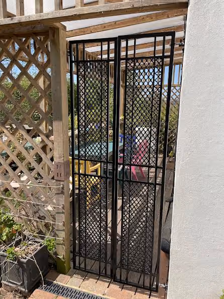 Metal gate with black lattice design partially open to a patio with colorful chairs.