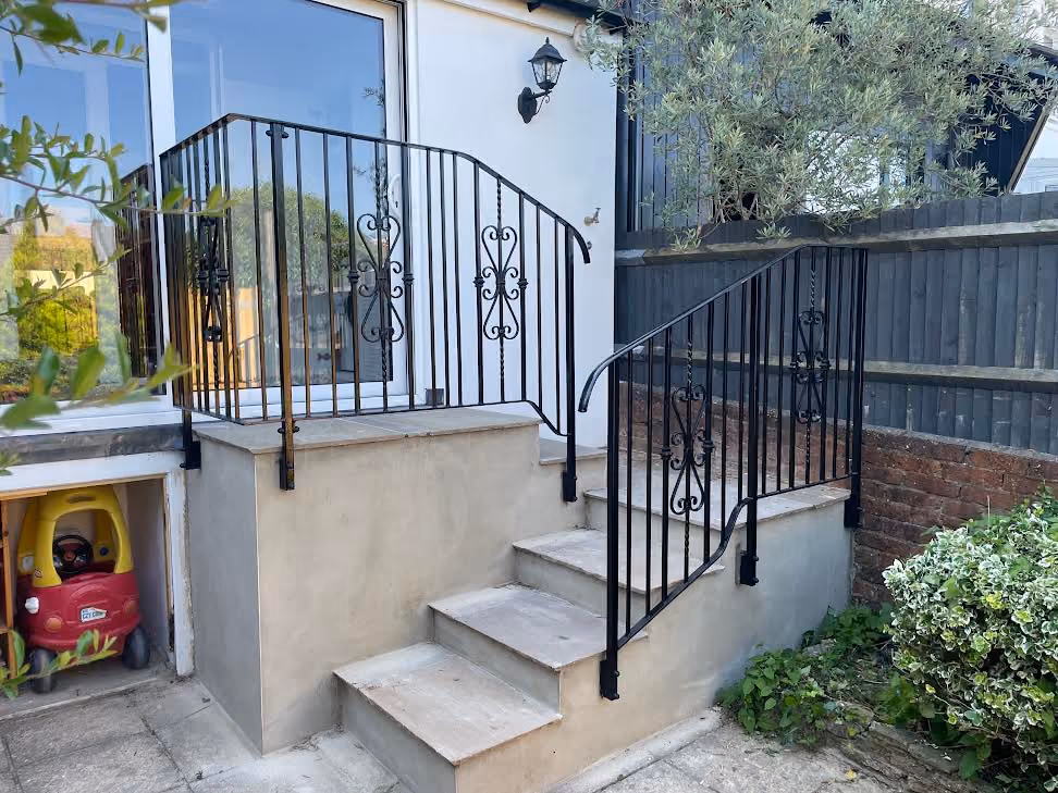 Outdoor steps with black iron railings leading to a door, surrounded by greenery.