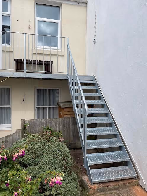 Metal stairs leading to a second-floor apartment door, with plants below in the small garden area.
