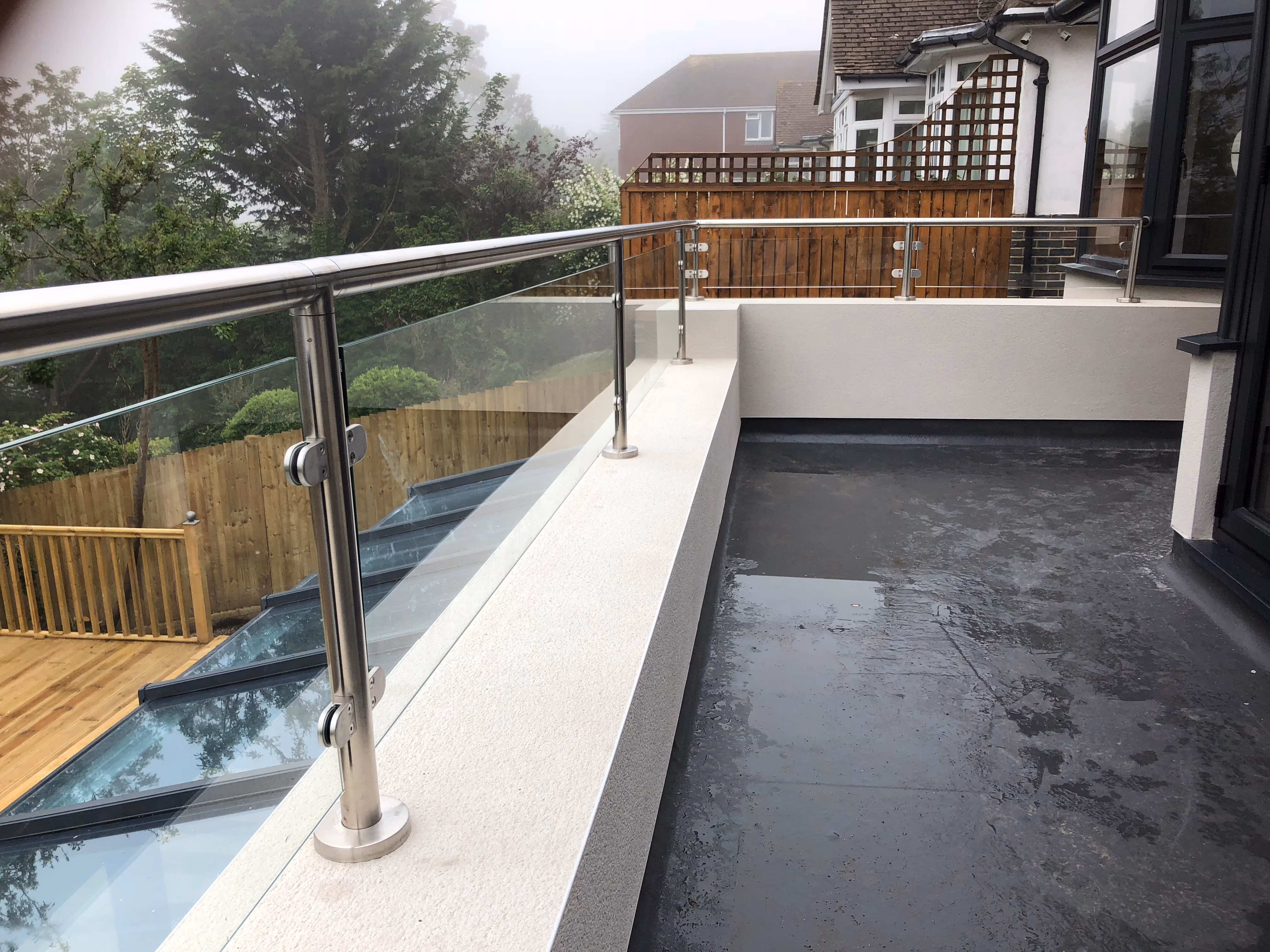A wet balcony with a glass railing overlooks a wooden deck and garden below.