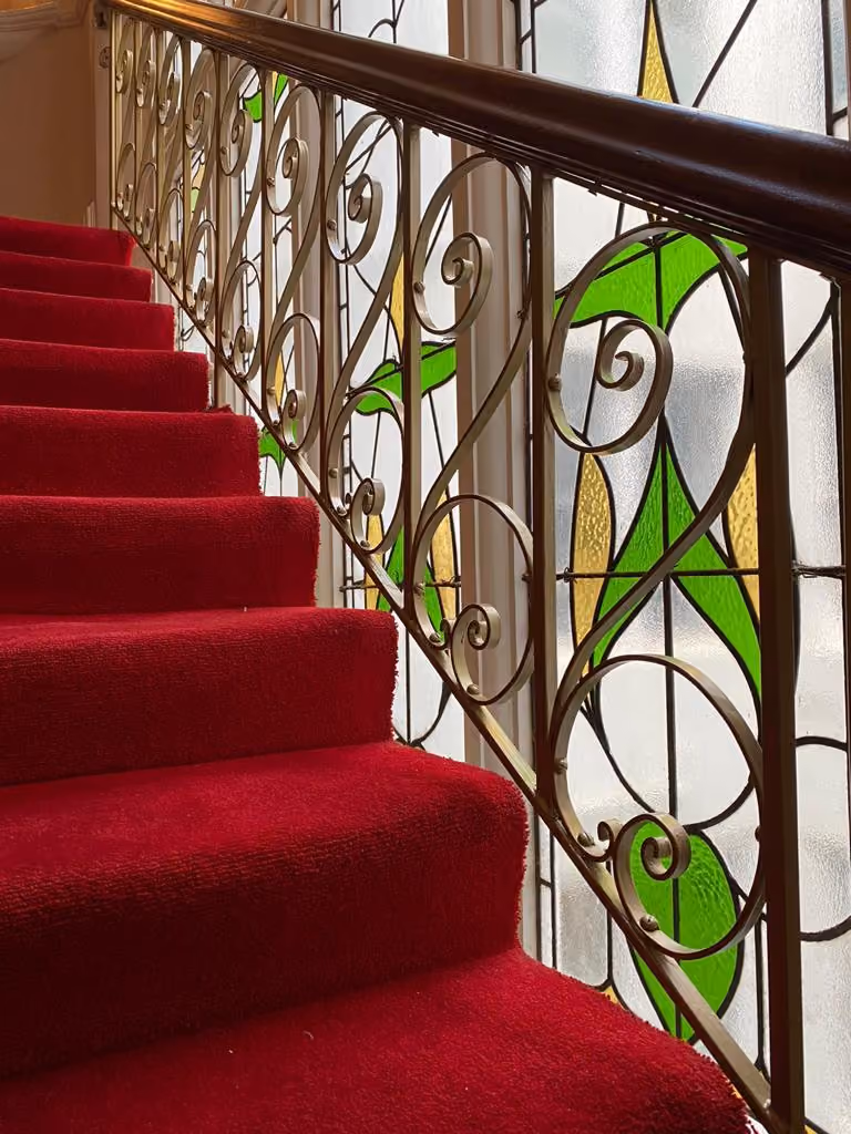 Red carpeted stairs beside ornate metal railing with stained glass window featuring green and yellow designs.