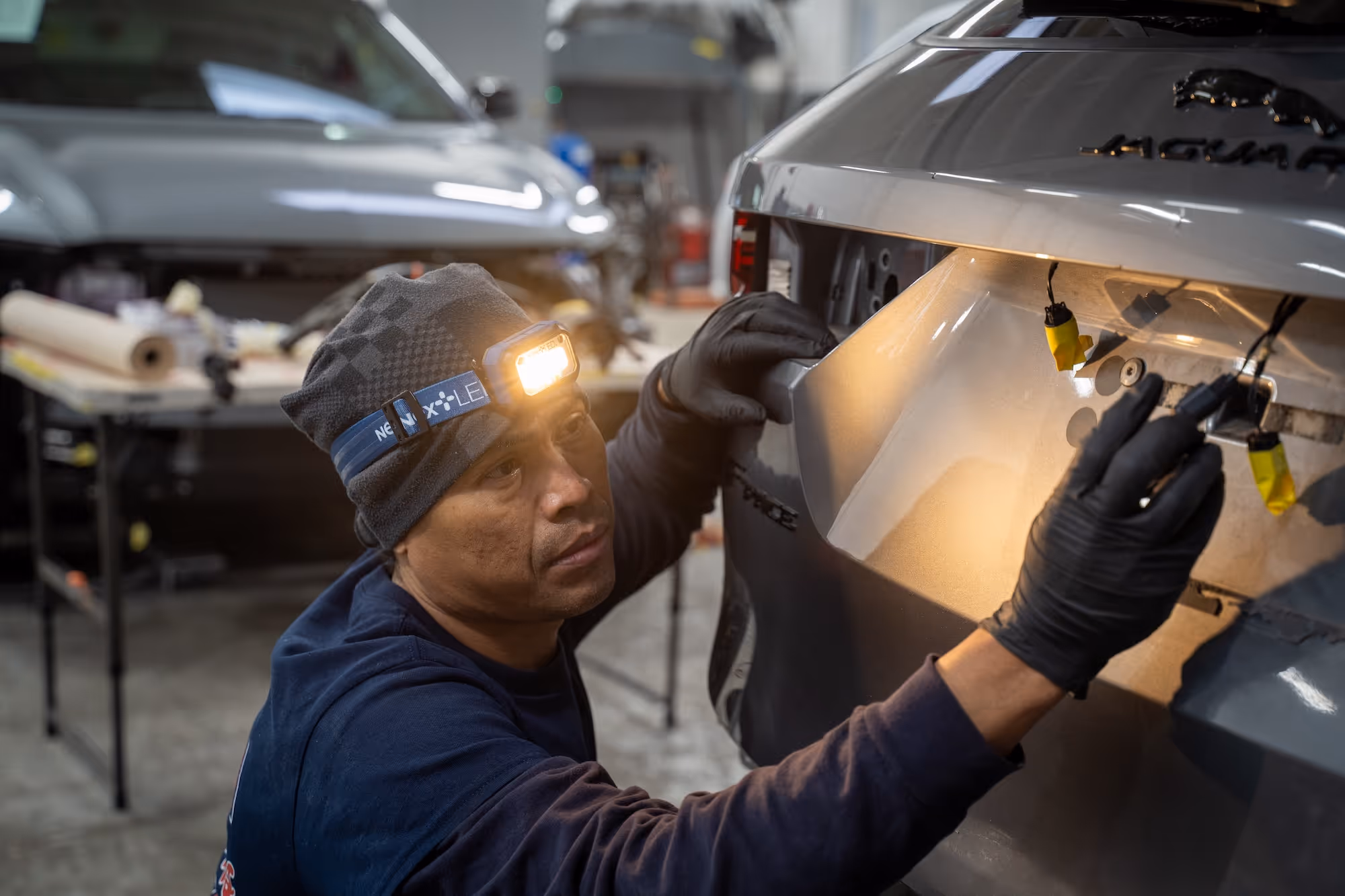 Auto body technician repairing the wiring of a grey Jaguar vehicle.