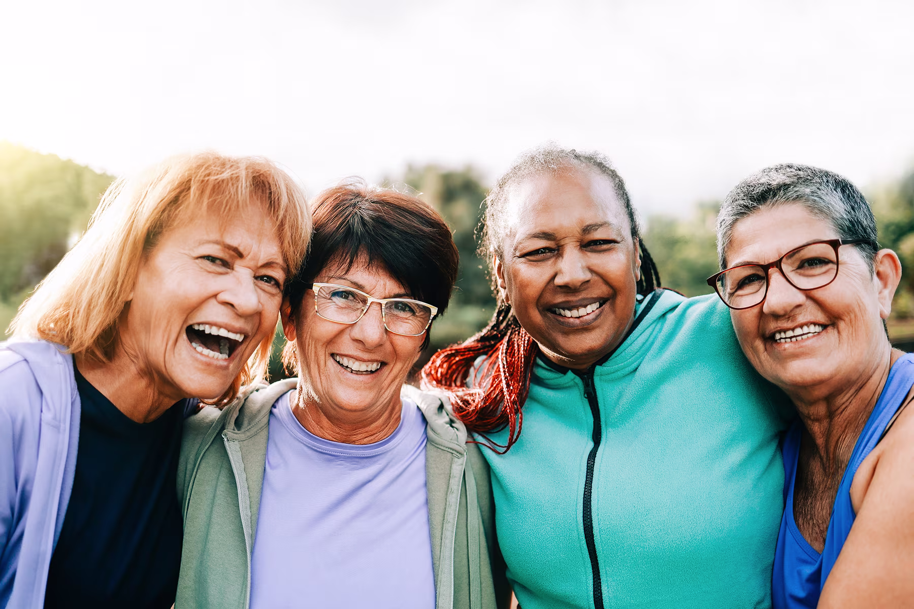 Four middle-aged women smiling cheerfully outdoors wearing casual athletic clothes.