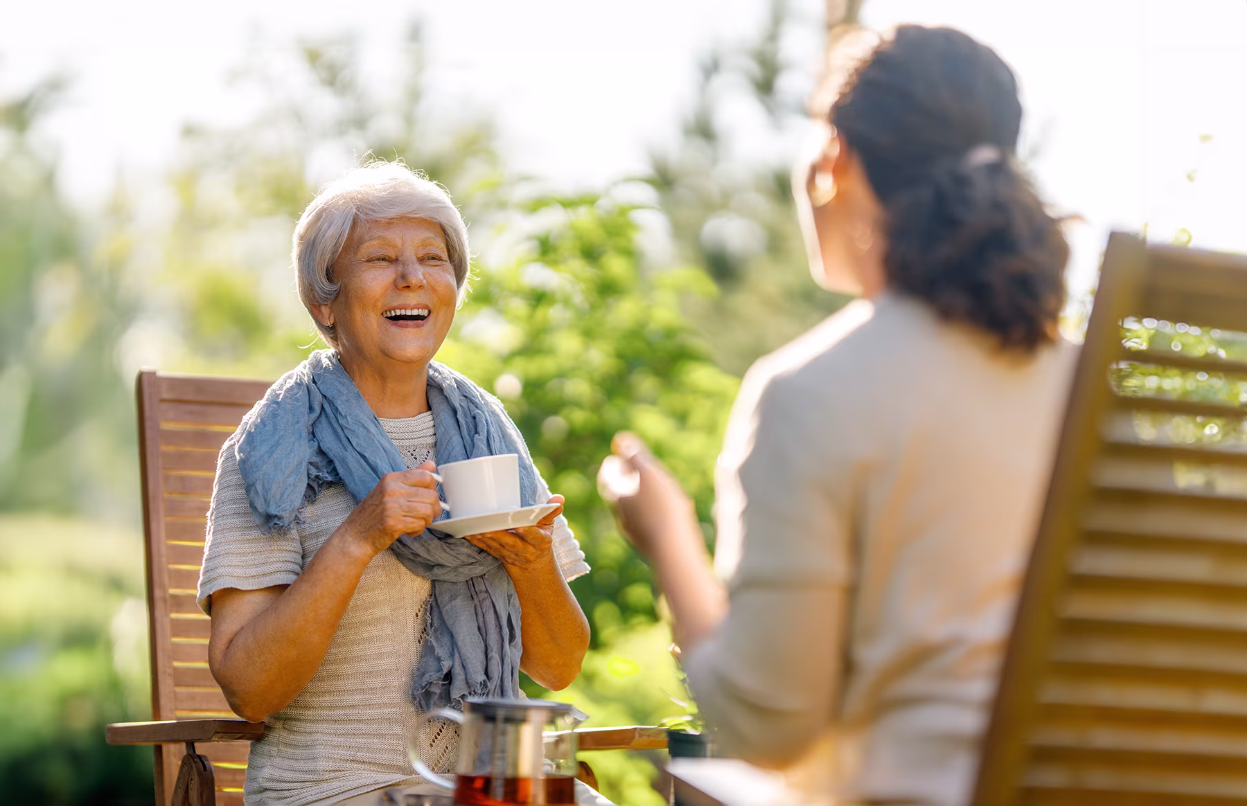 Older woman with gray hair smiling and holding a teacup while sitting outdoors with a younger woman.