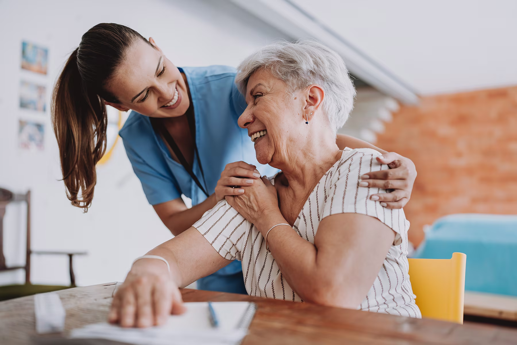 Smiling caregiver in scrubs embraces cheerful elderly woman sitting at a table in a home setting.