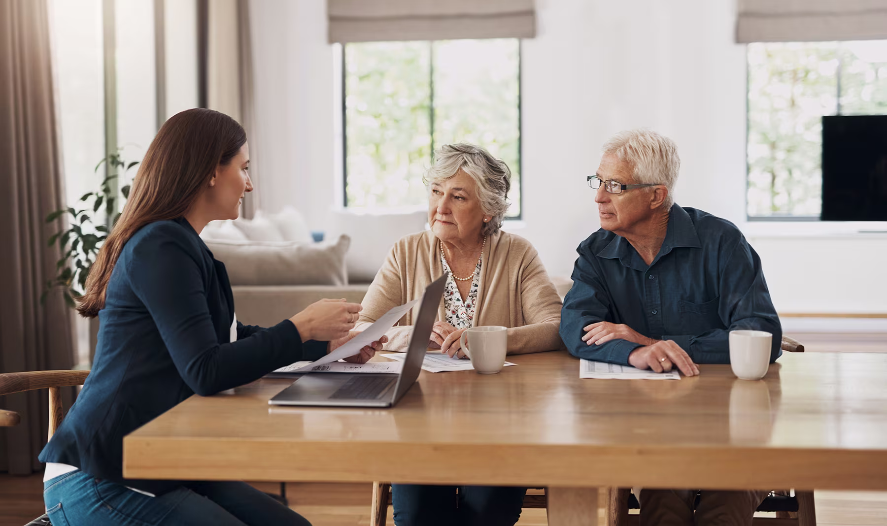Young woman discussing documents with an elderly couple at a wooden table in a bright room.