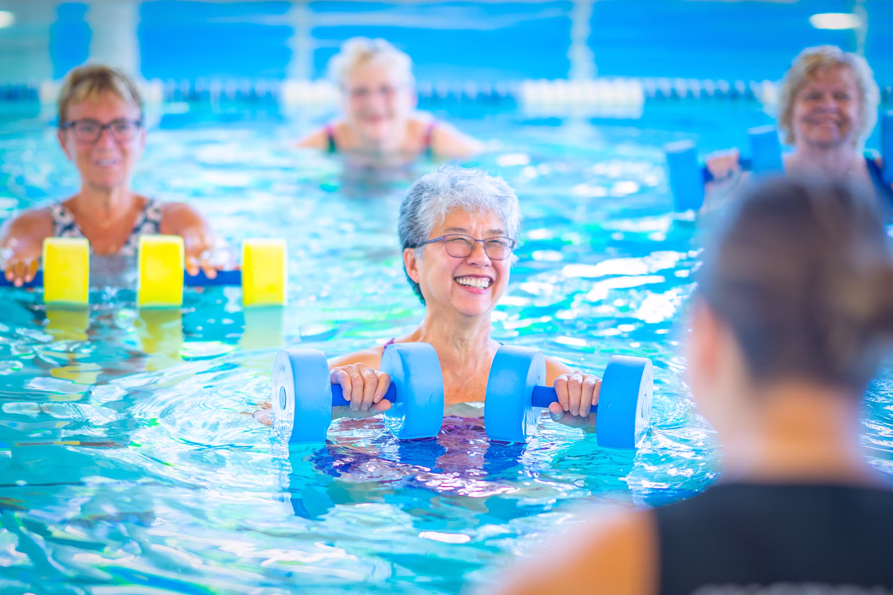 Smiling older women exercising with foam dumbbells in a pool during a water fitness class.